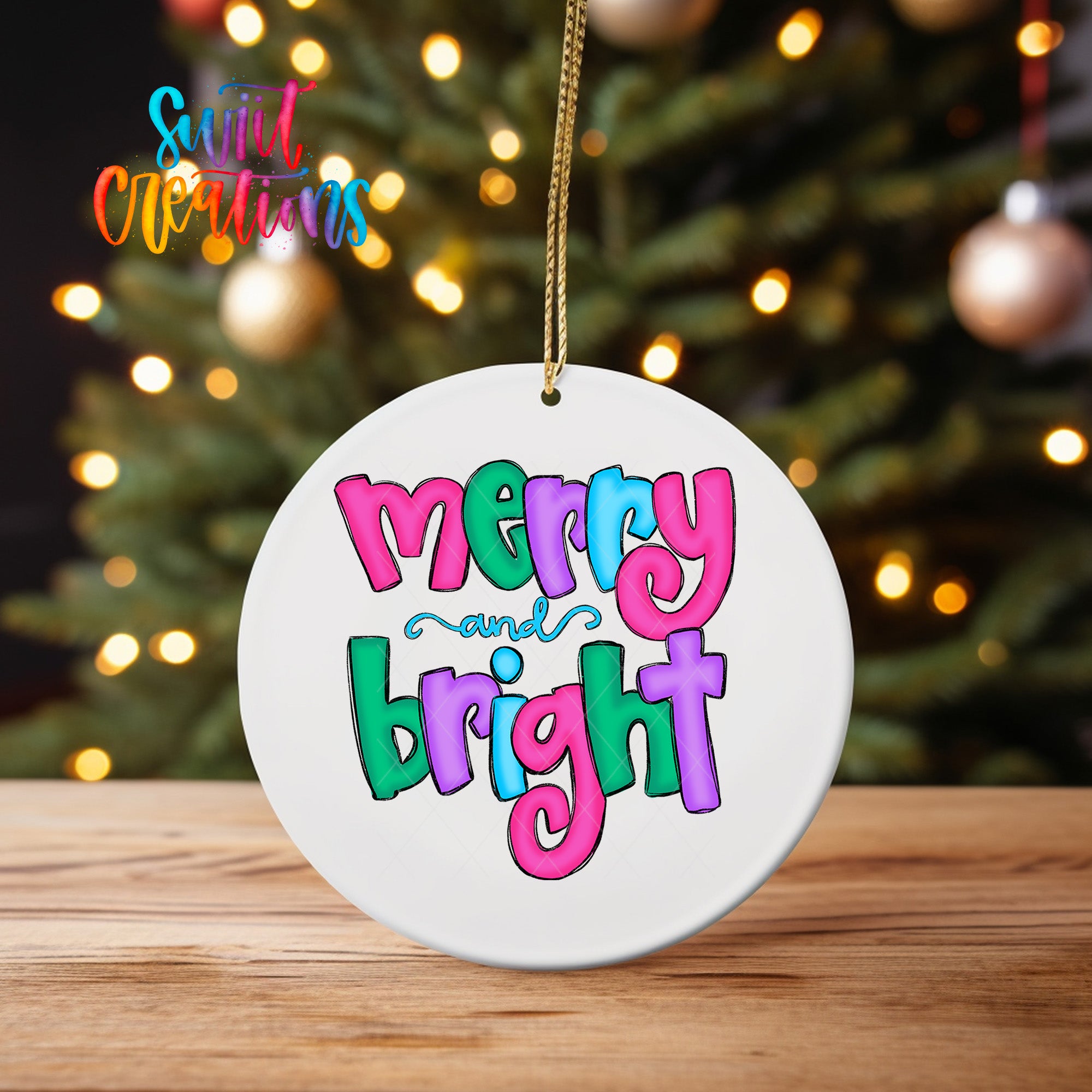 A white ornament with the text "mercy and bright" in colorful letters hangs from a string against a blurred background of a Christmas tree with twinkling lights.