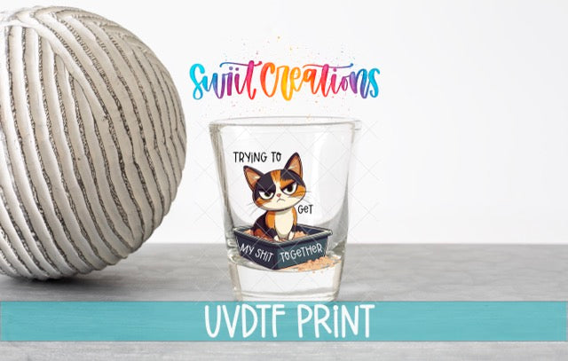 a glass with a cat illustration and text, placed next to a white ball on a gray surface.