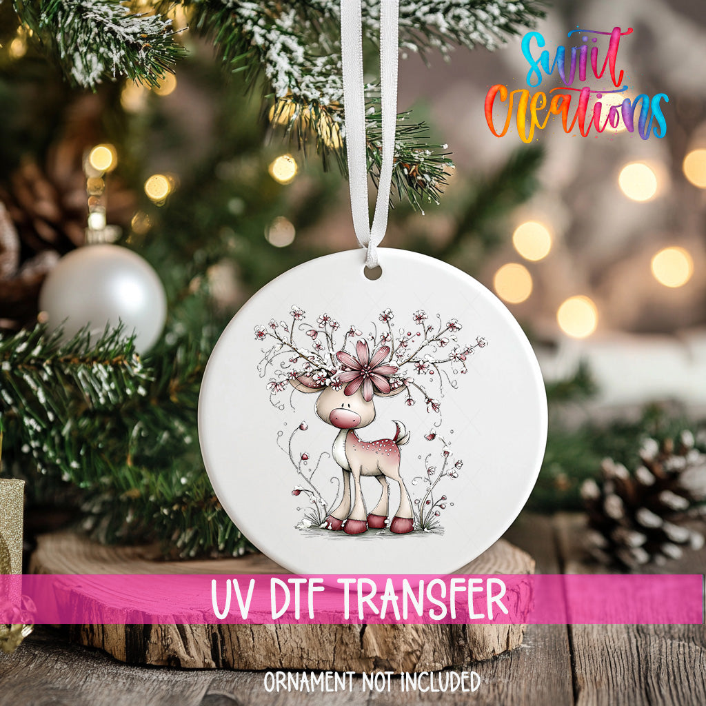 A white ceramic ornament with a deer and floral design hangs on a Christmas tree, with a blurred background of lights and greenery.