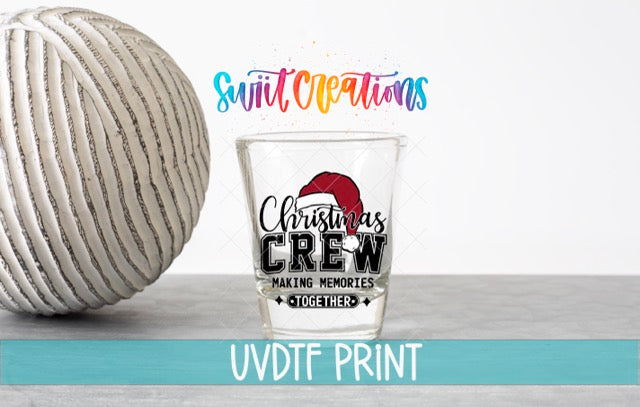 a shot glass with a Christmas crew design and text, along with a white ball and a blue banner with text.