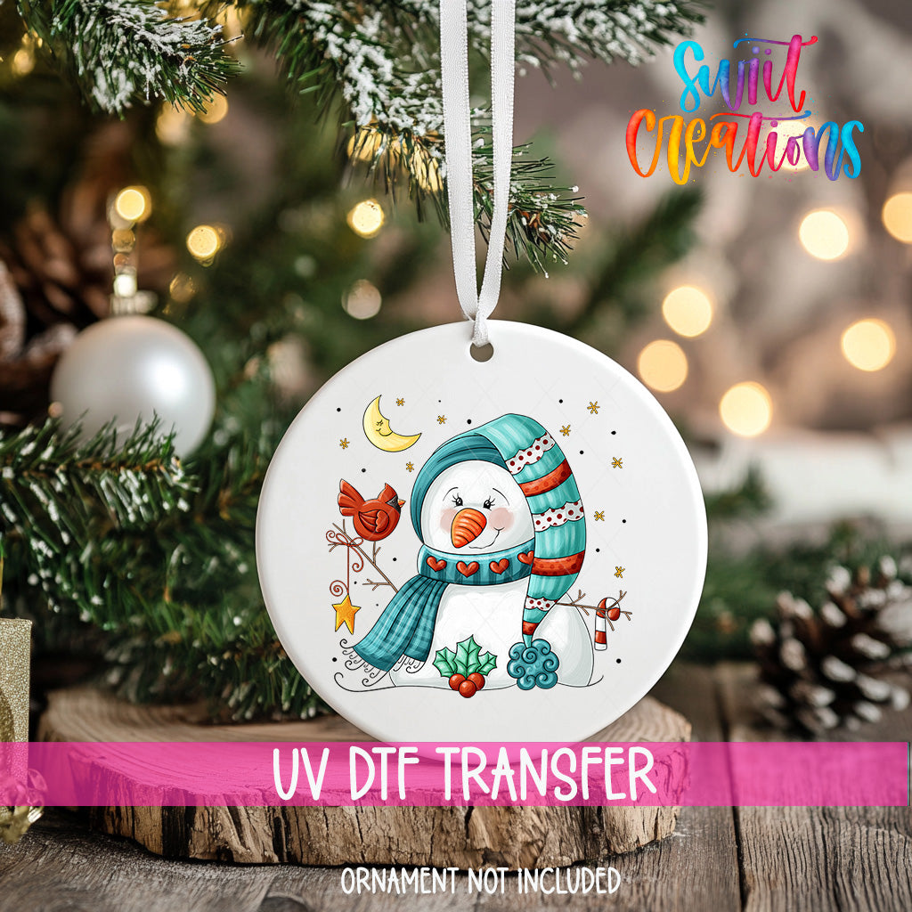 A white ceramic ornament with a snowman design, featuring a scarf, hat, and other winter-themed elements, hangs on a Christmas tree.