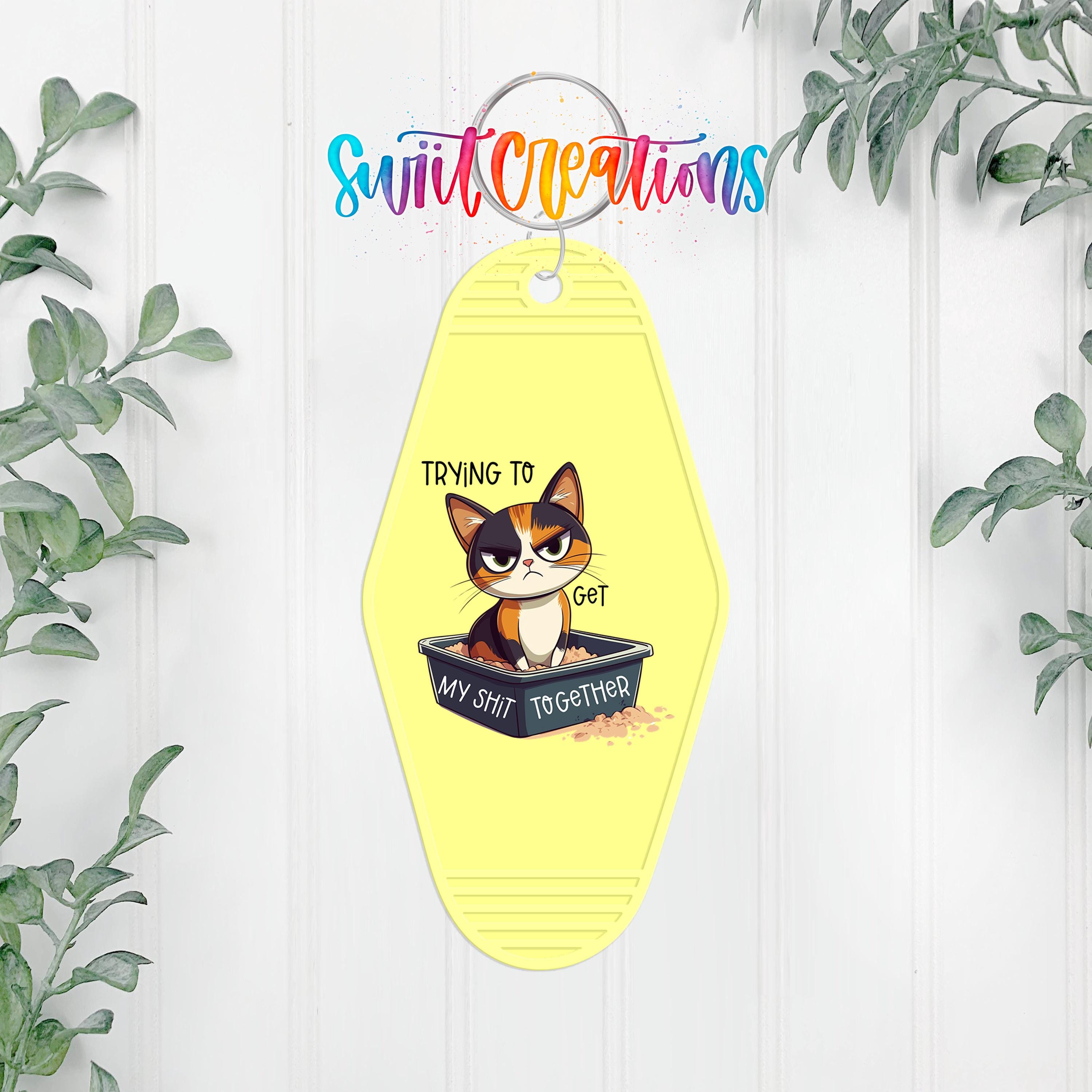 A yellow keychain with a cartoon cat and the text "Trying to get my shit together" hanging on a wall.