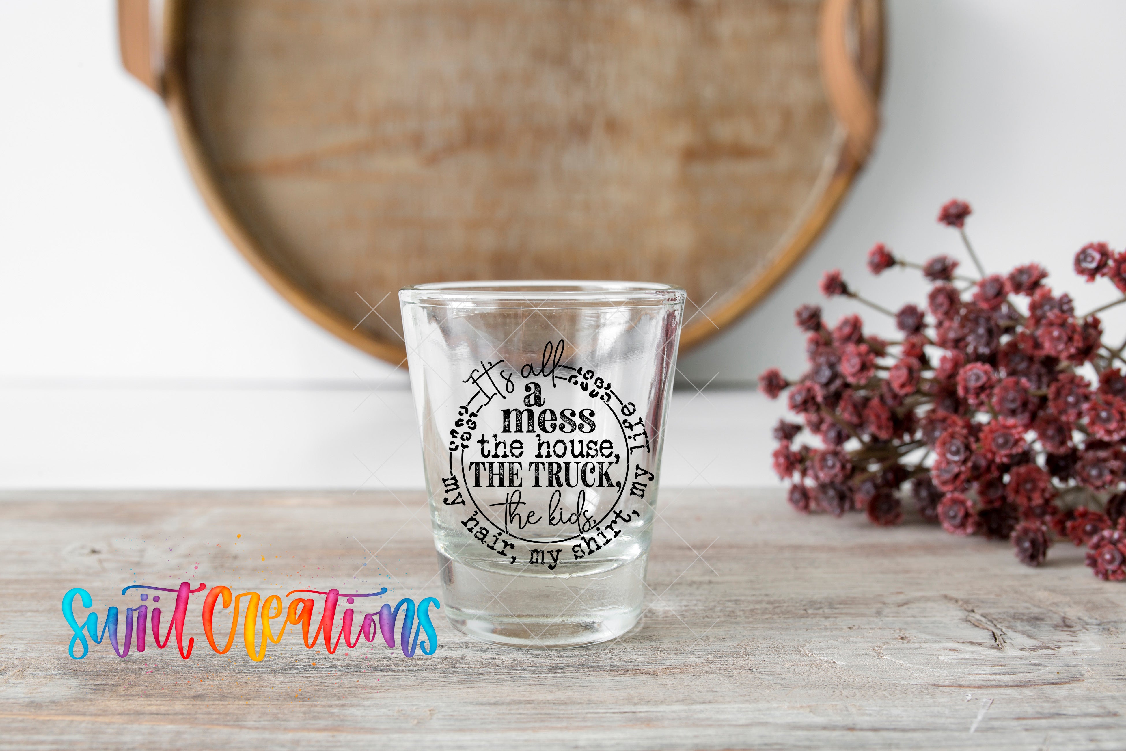 A shot glass with a printed message on it, placed on a wooden surface next to a bunch of red flowers.
