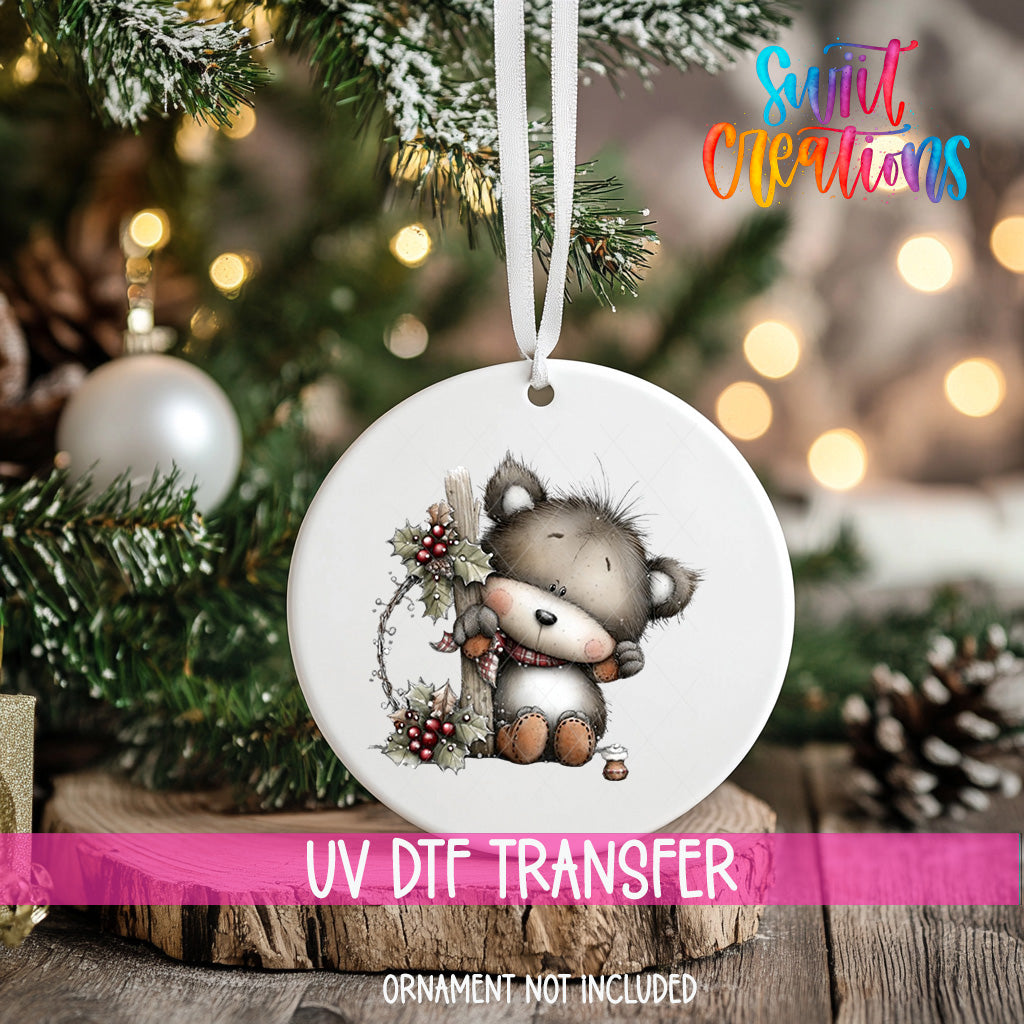 A white ornament with a cute bear design hangs on a Christmas tree, surrounded by pine branches and other holiday decorations.