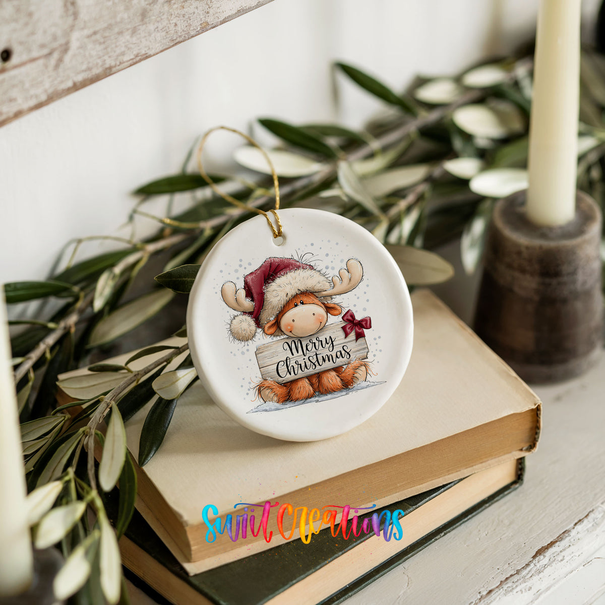 moose in a Santa hat holding a Merry Christmas sign ceramic Christmas ornament styled on stacked books with greenery