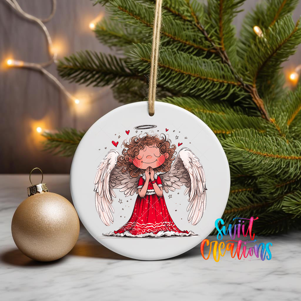A white ceramic ornament featuring an illustration of a smiling angel with curly hair and wings, surrounded by hearts and flowers, is displayed on a table next to a gold ornament and a Christmas tree.