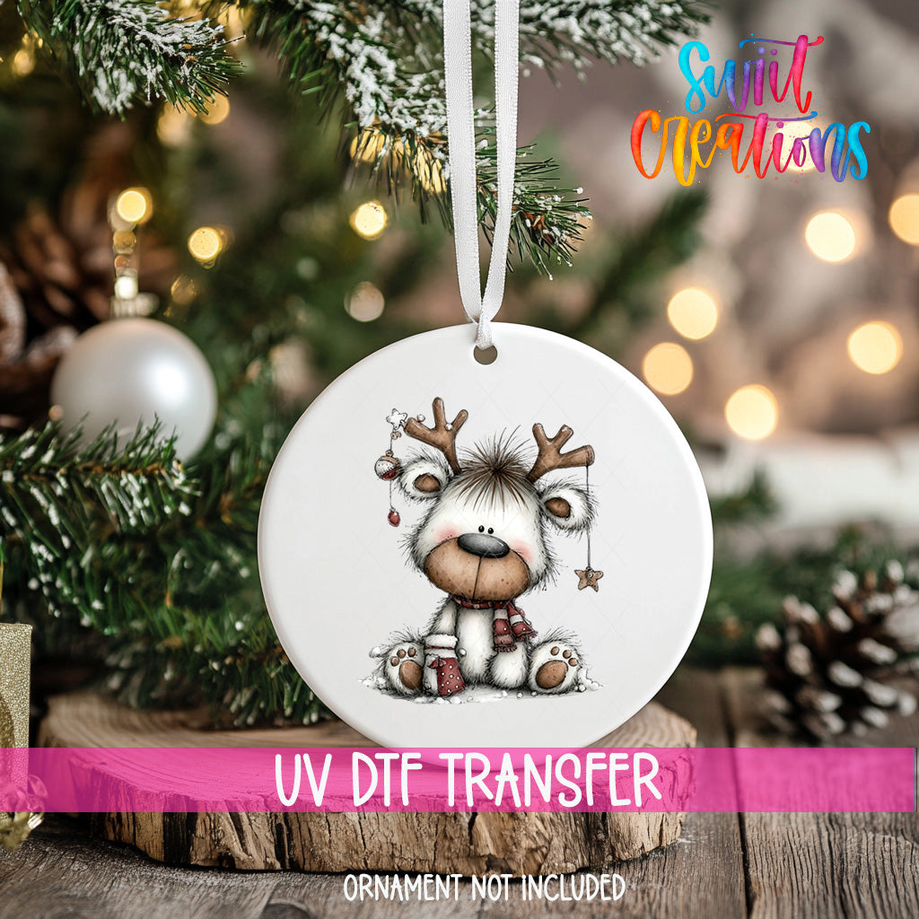 A white ornament with a cute reindeer design hangs on a Christmas tree, surrounded by pine branches and twinkling lights.