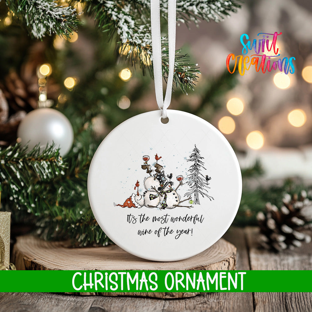 A white Christmas ornament with a snowman and pine trees, hanging on a tree branch.