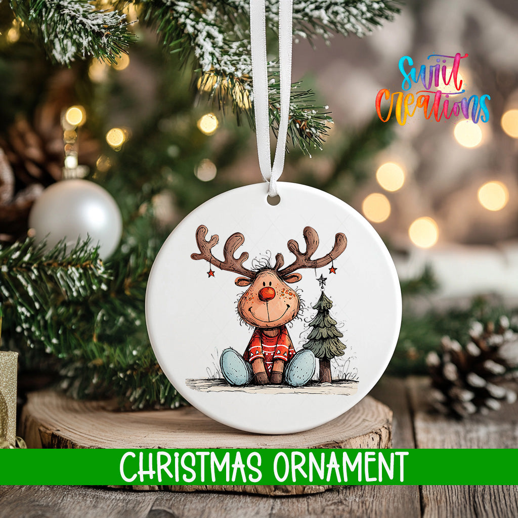 A white Christmas ornament featuring a cartoon reindeer sitting on a Christmas tree, with a tree branch and pine cones in the background.