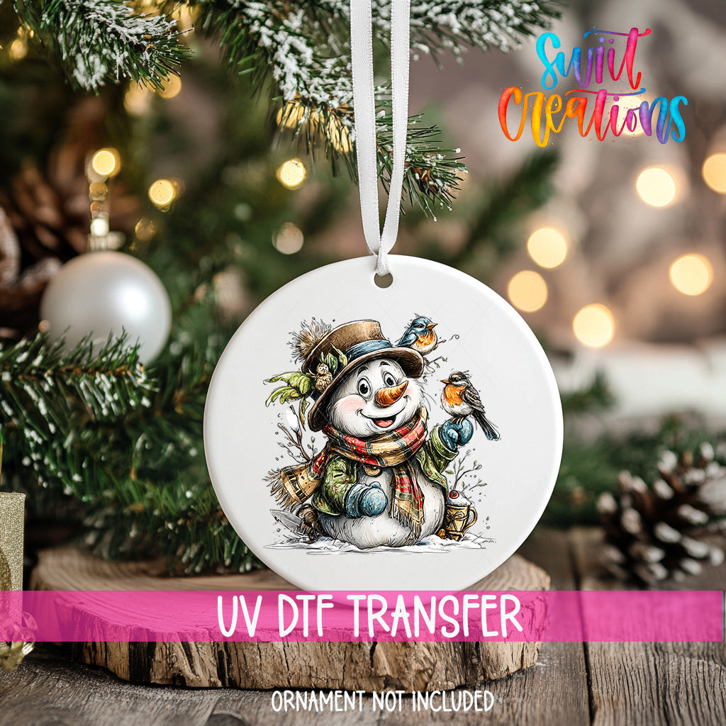A white ornament with a snowman and a bird is hanging on a Christmas tree, with a pink ribbon around it.