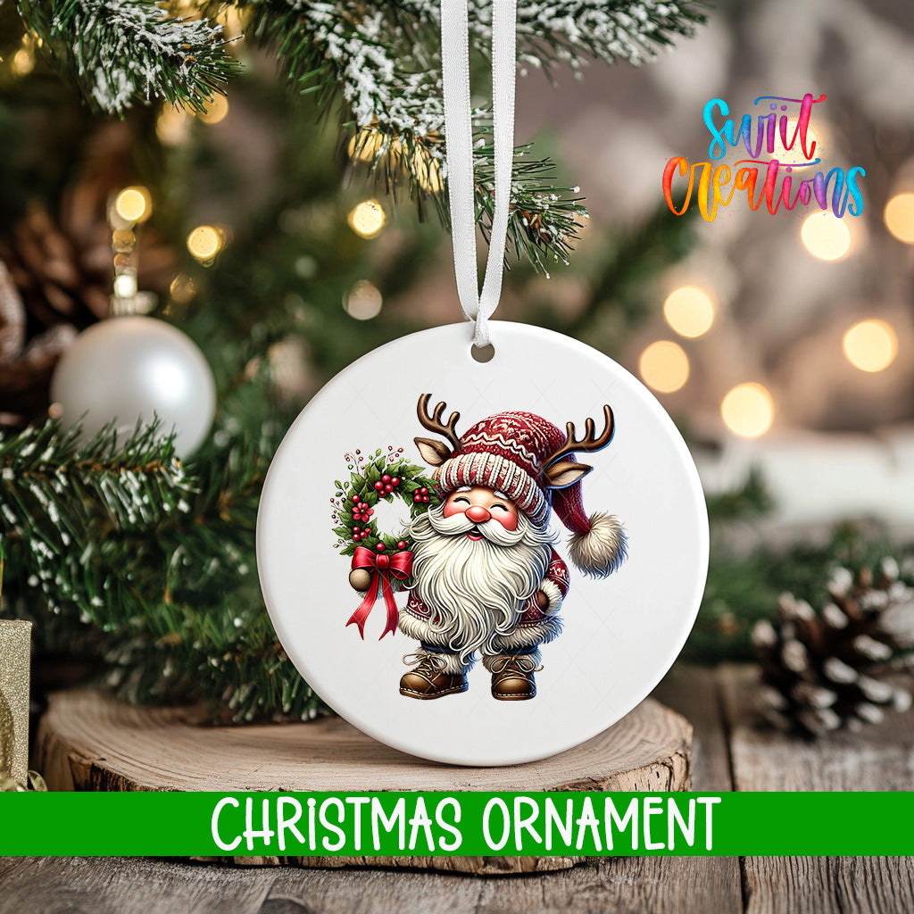 A white Christmas ornament featuring a Santa Claus figure holding a wreath and a bow tie, hanging on a tree branch with a blurred background of lights and greenery.