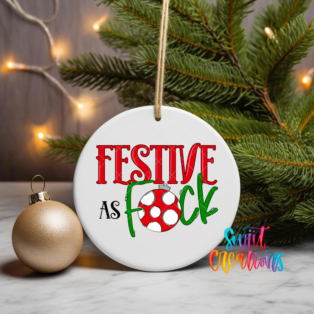 A white ornament with the text "FESTIVE AS FOCK" in red and green colors, along with a red and white polka dot design, is hanging on a Christmas tree.