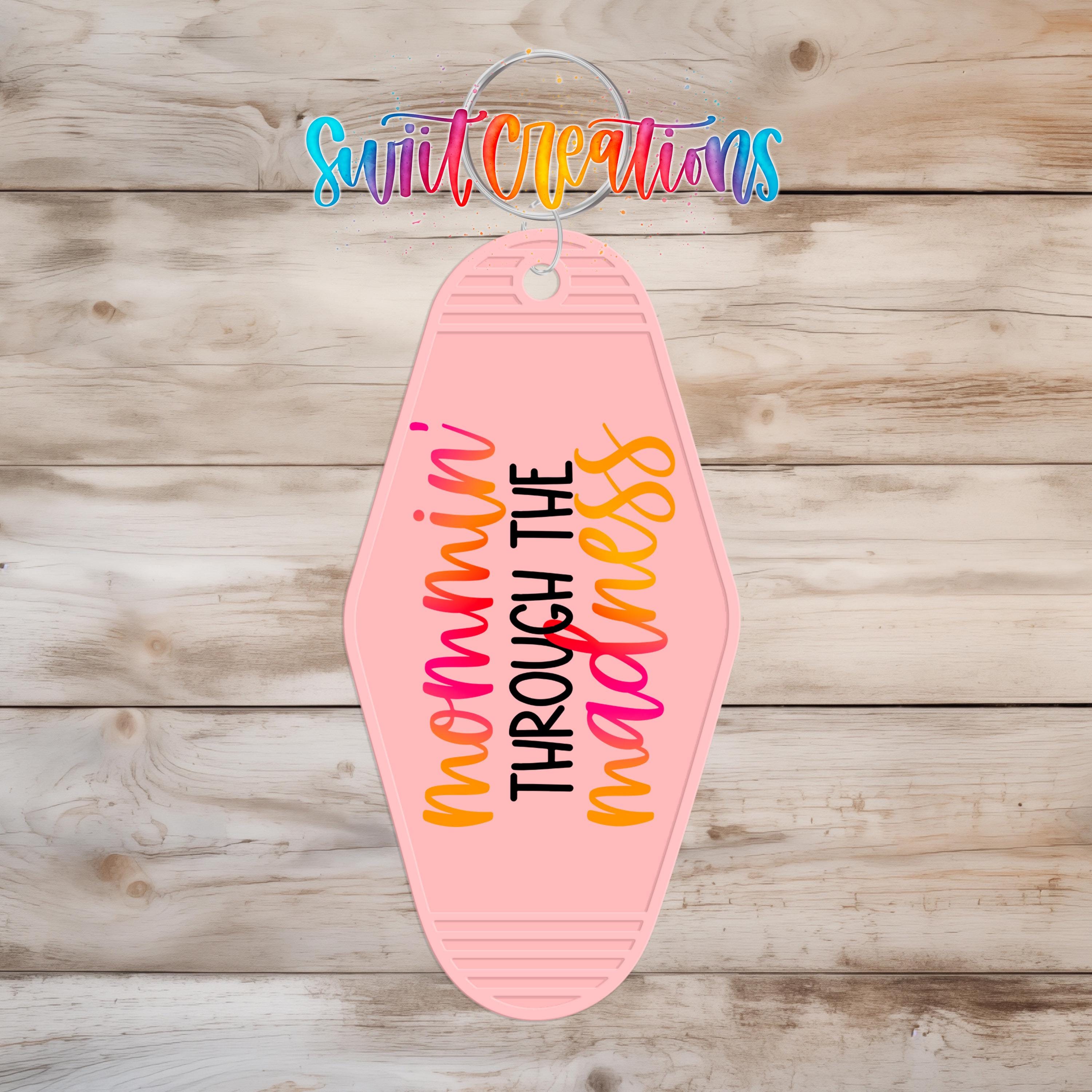 A pink keychain with the text "nommini through the madness" printed on it, hanging on a wooden surface.