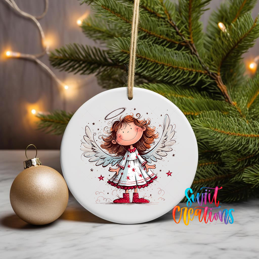 A white ceramic ornament featuring a smiling cartoon girl with wings, surrounded by stars and a halo, is displayed on a table next to a gold ornament and a Christmas tree.