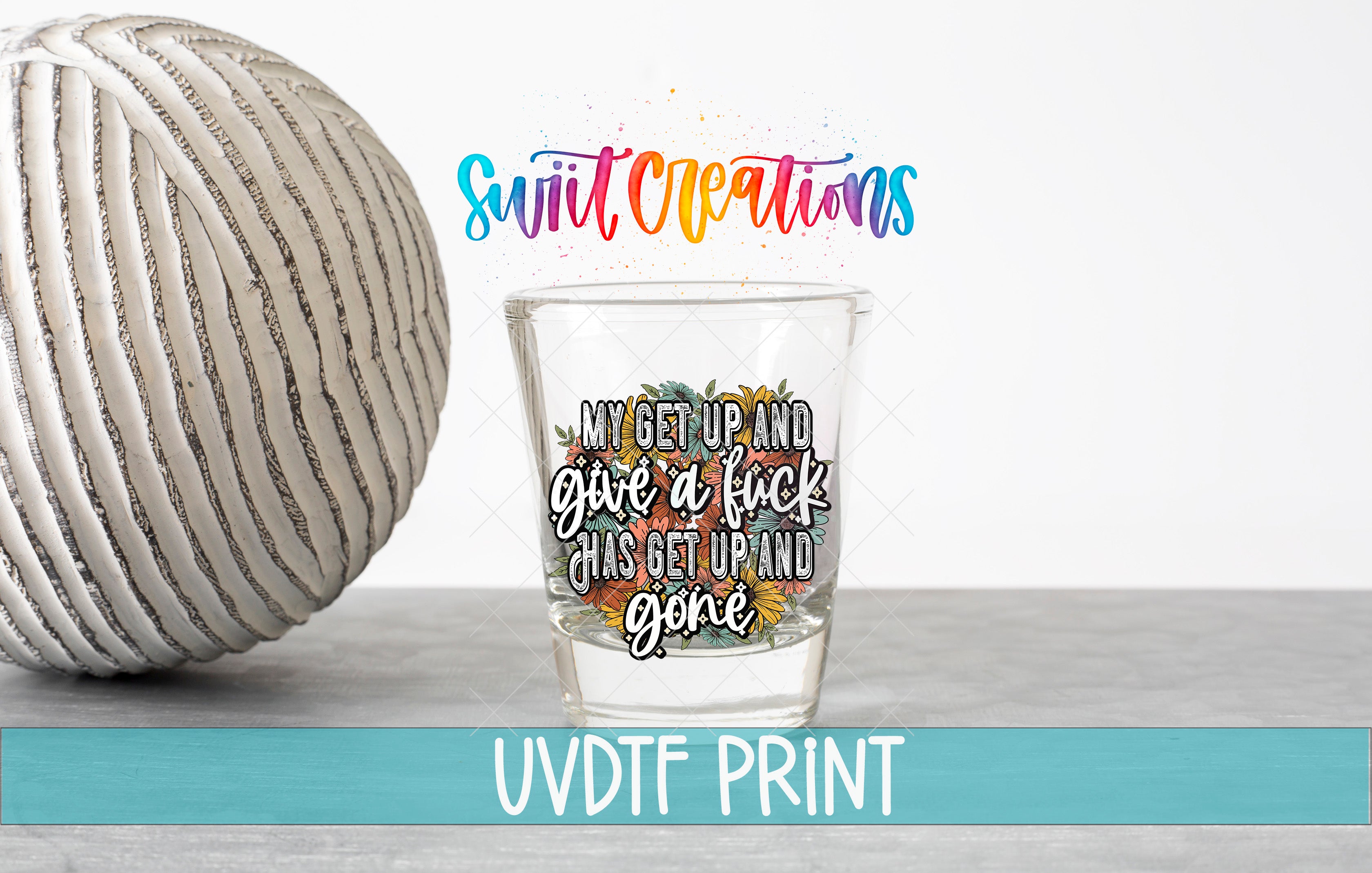 a shot glass with a colorful design and text, placed next to a white ball on a gray surface.