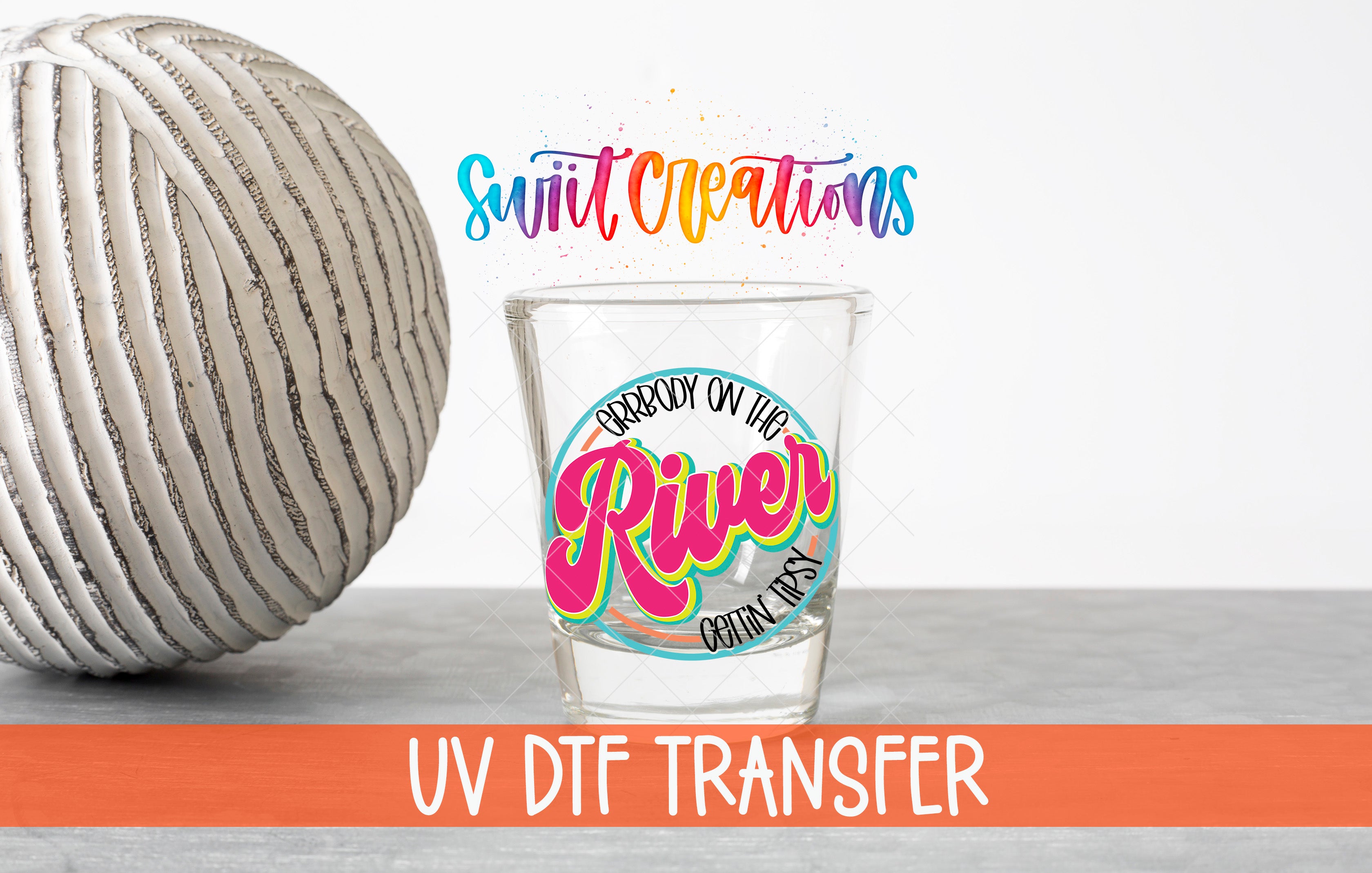 a glass with a colorful design and text, placed next to a white ball with a striped pattern. The text on the glass reads "Swift Creations" and "UV DFT Transfer".