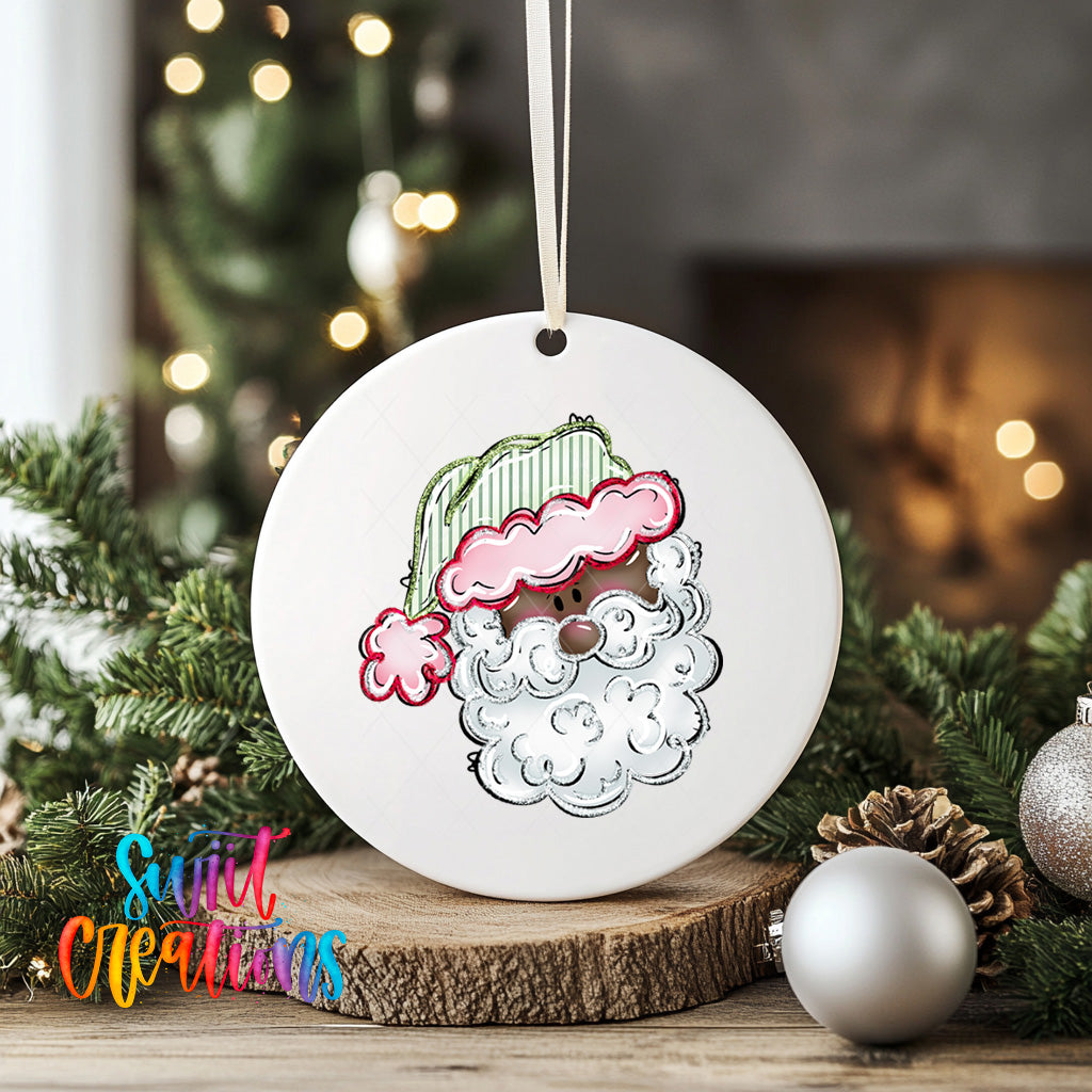 A white ceramic ornament with a Santa Claus face design hangs on a wooden surface, surrounded by Christmas decorations and greenery.