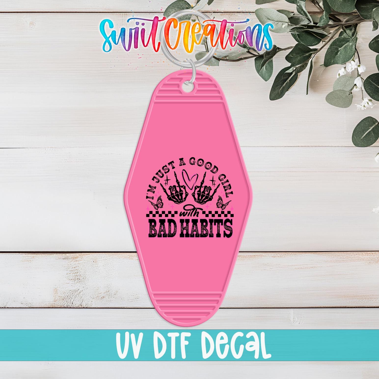 A pink keychain with a black and white graphic design and text, placed on a wooden surface with greenery in the background.