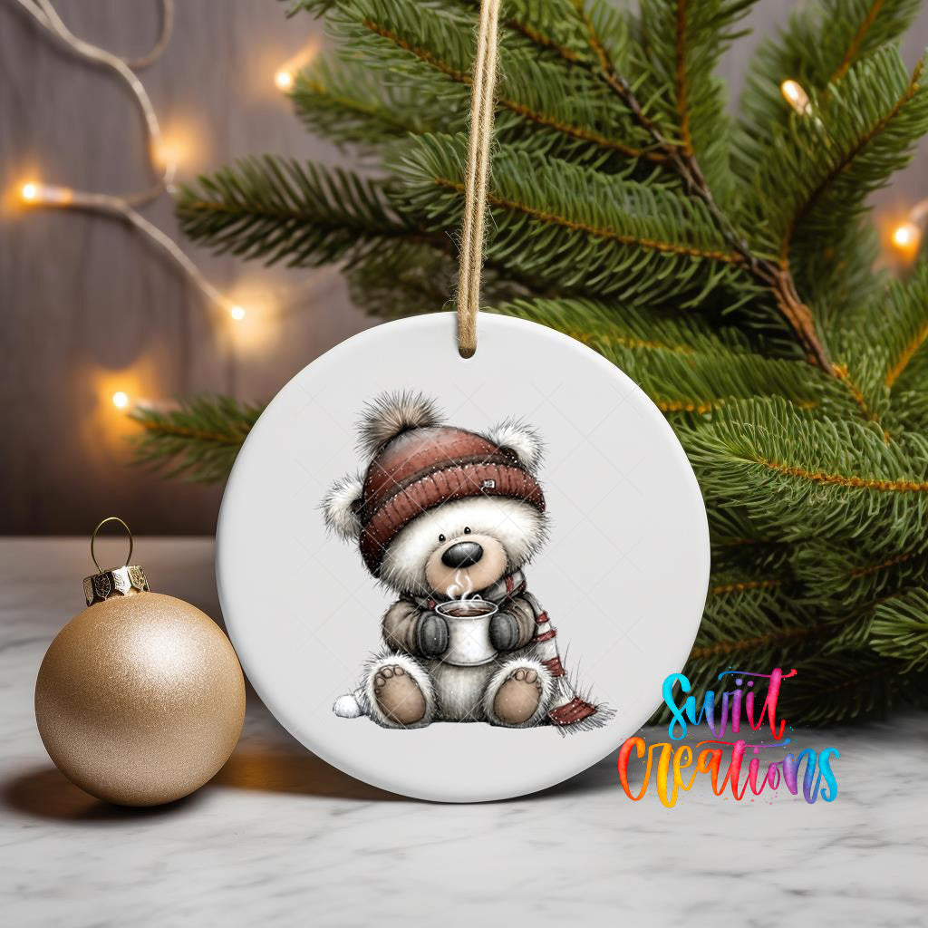 A white ceramic ornament featuring a teddy bear wearing a red hat and scarf, sitting next to a gold ornament and a Christmas tree with twinkling lights.