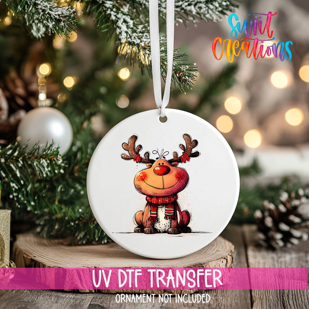 A white ornament with a cartoon reindeer design hangs on a Christmas tree, surrounded by pine branches and twinkling lights.