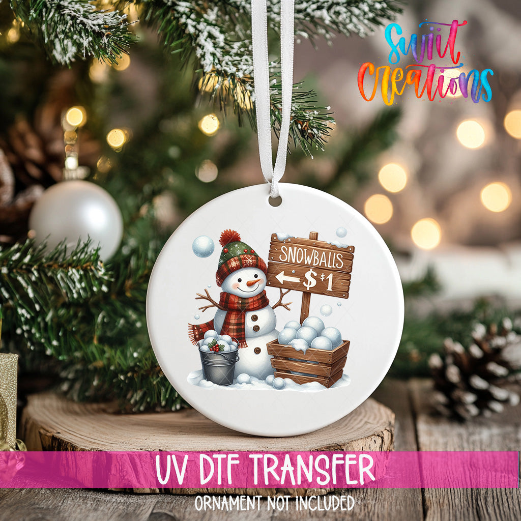 A white ornament with a snowman and a sign that says "Snowballs $1" hanging on a Christmas tree.