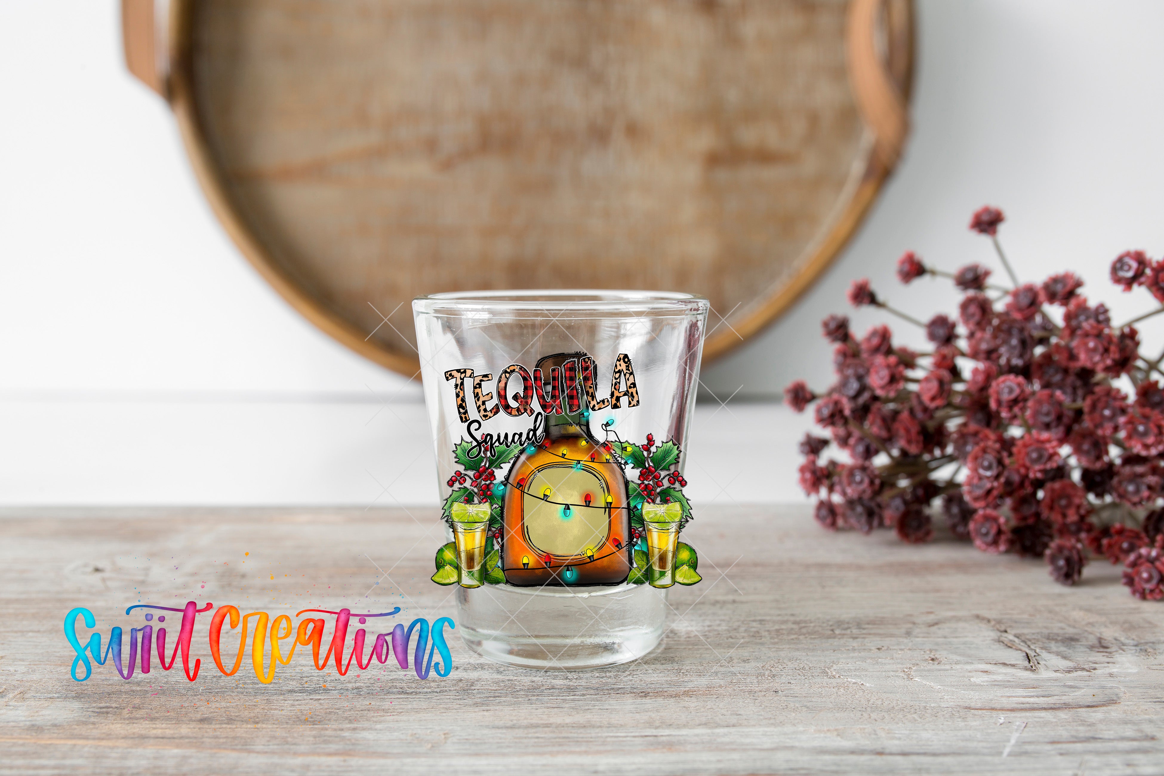 A clear shot glass with a tequila-themed design, featuring a bottle of tequila, a lime, and a tequila shot glass, placed on a wooden surface.
