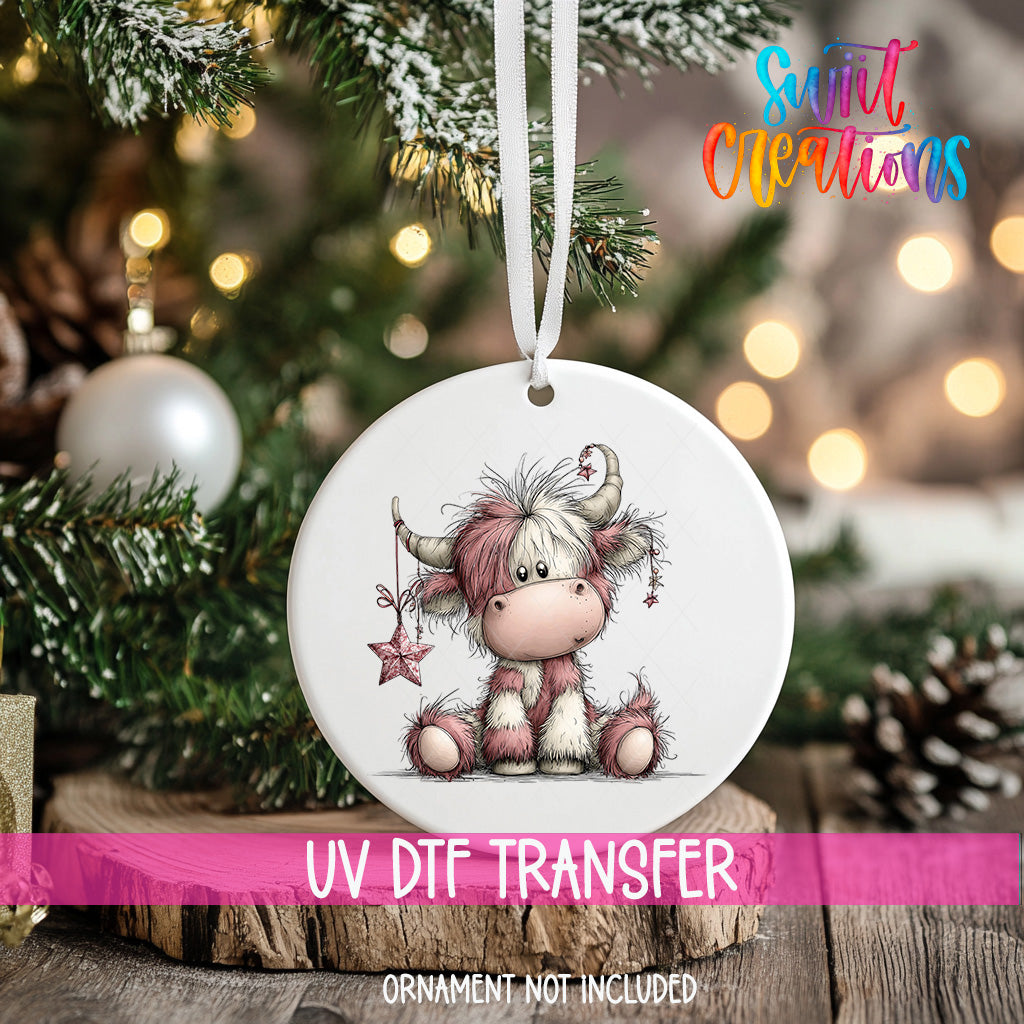 A white ceramic ornament with a cartoon cow design hangs on a Christmas tree, surrounded by pine branches and twinkling lights.