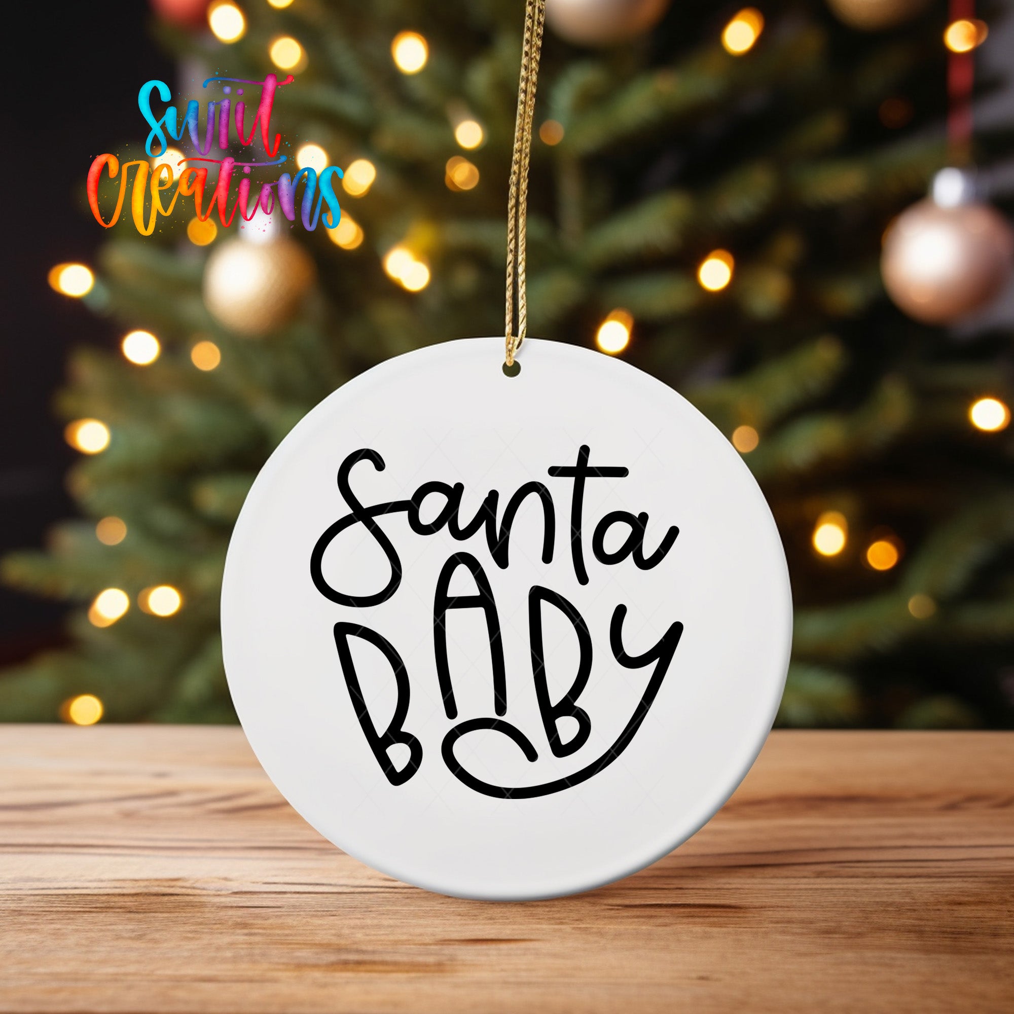 A white ornament with the text "Santa Baby" written on it, hanging from a string in front of a blurred Christmas tree with colorful lights.