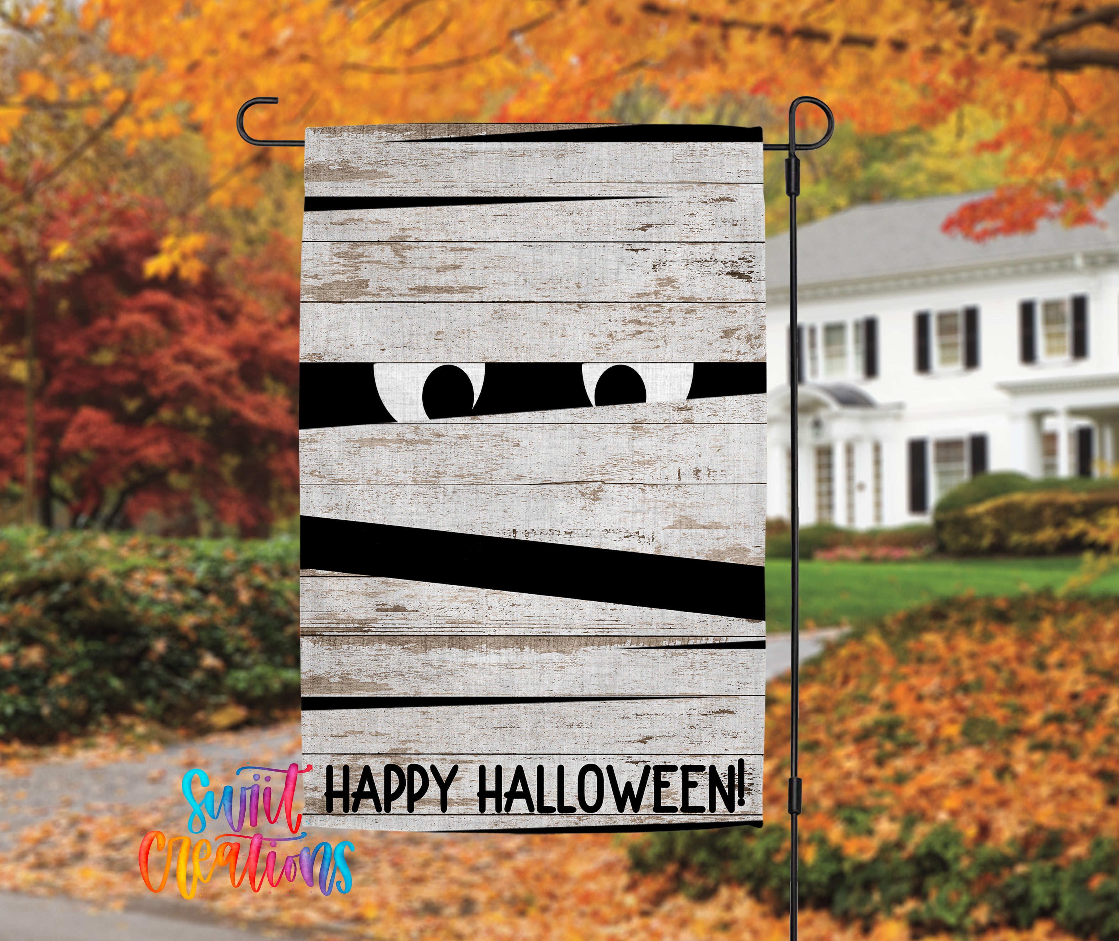 A Halloween-themed garden decoration with a wooden board displaying a mummified face and the text "Happy Halloween!" hanging on a metal pole.