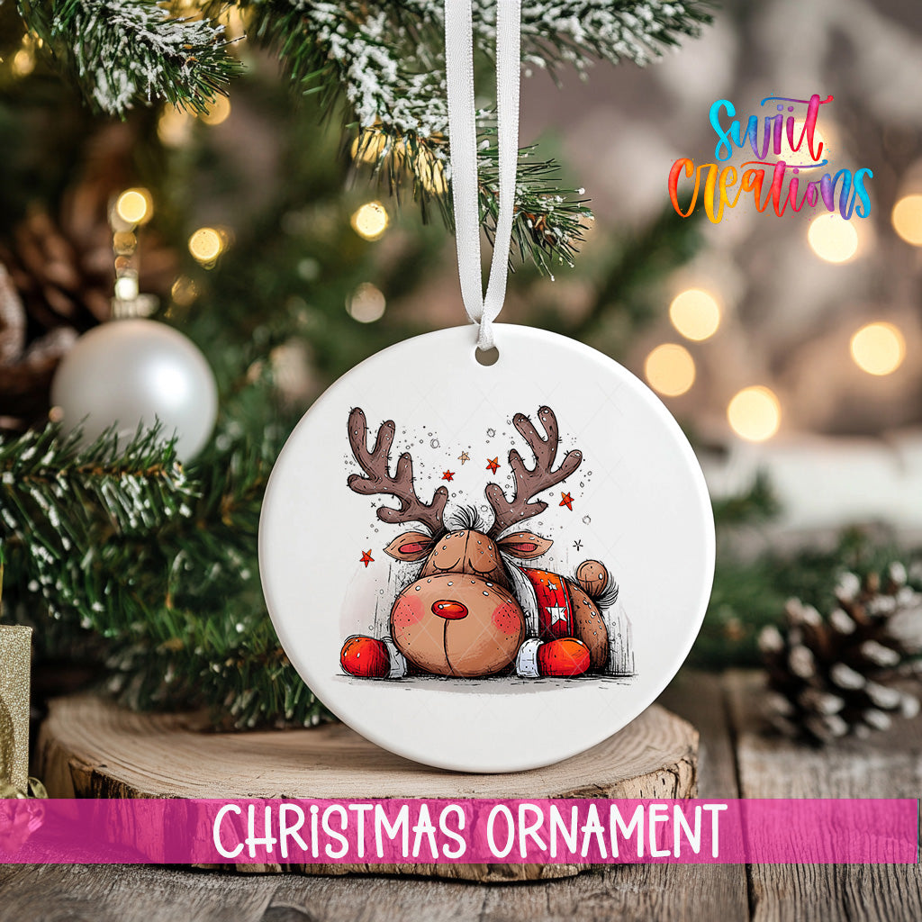 A white Christmas ornament featuring a cartoon reindeer is hanging from a tree branch, with a blurred background of lights and pine cones.