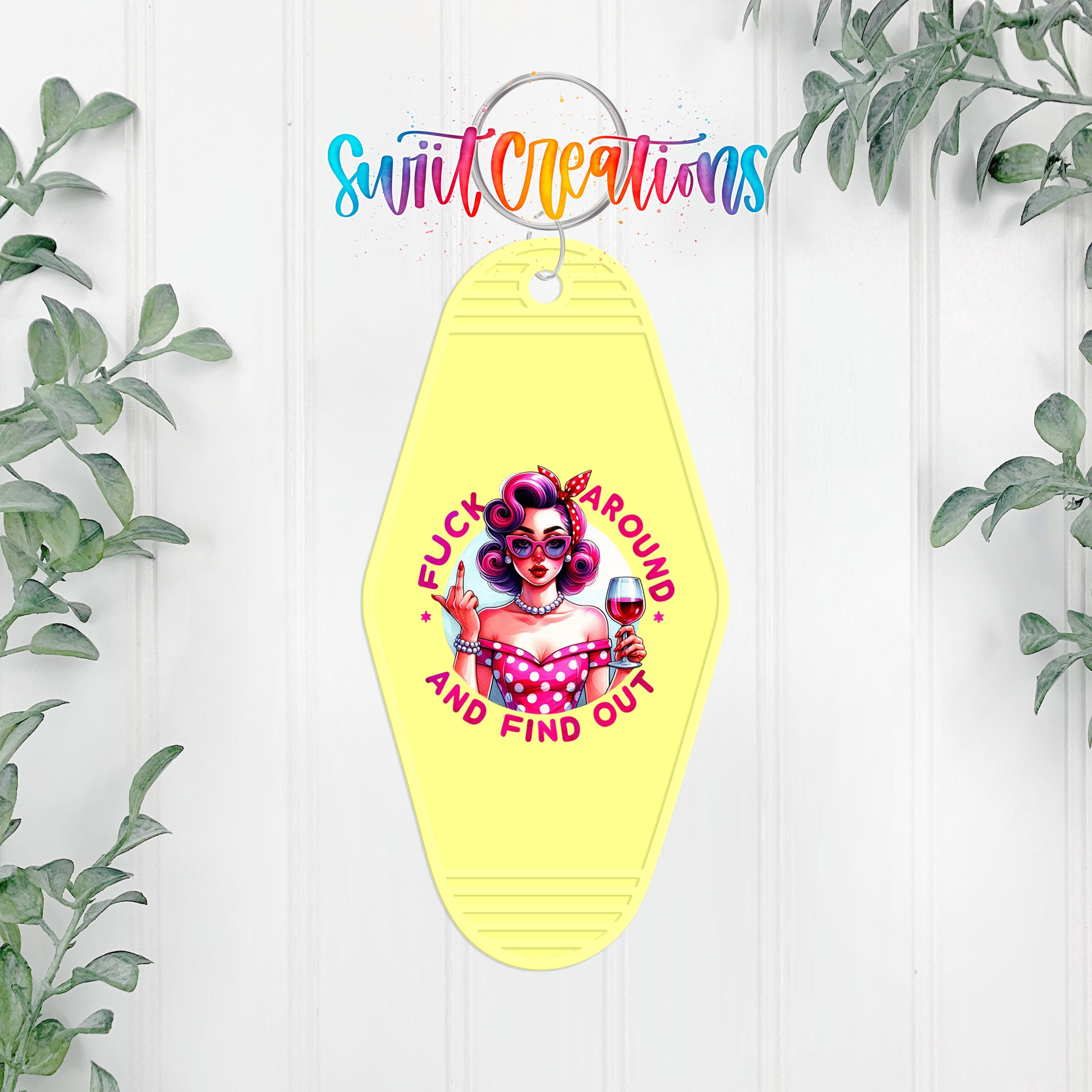 A yellow keychain with a graphic design featuring a woman with curly hair and a pink dress, along with the text "FUCK AROUND AND FIND OUT".
