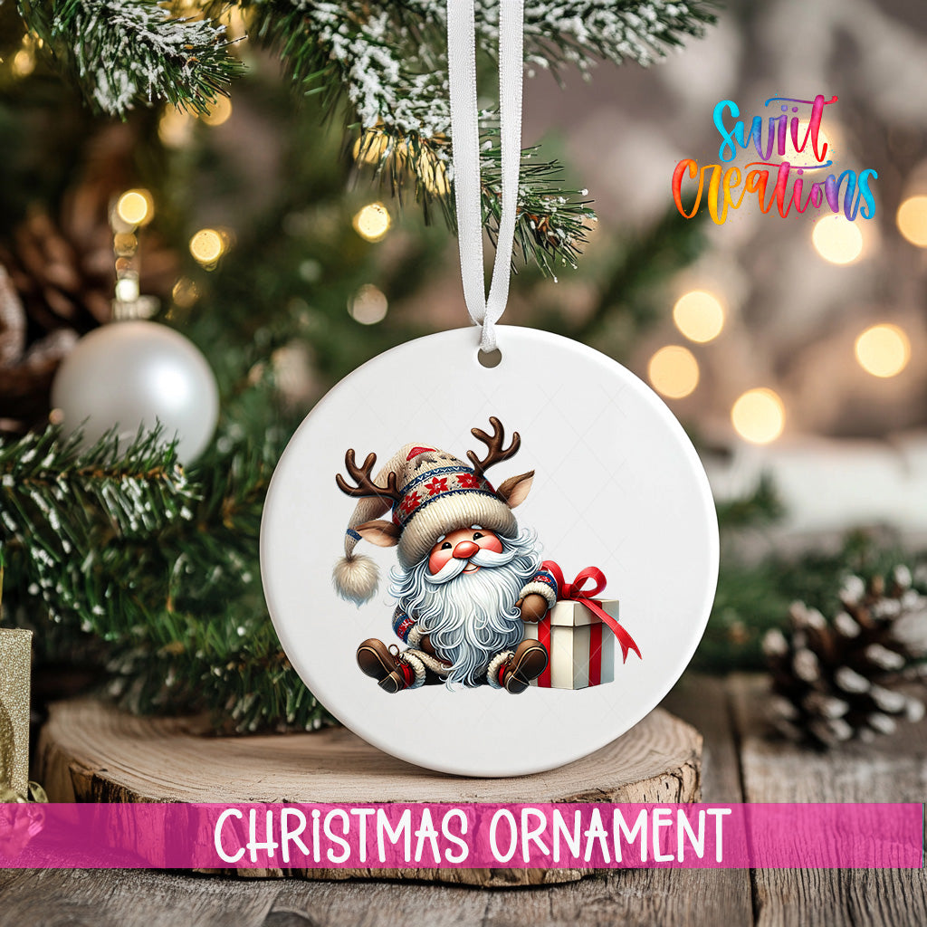 A white Christmas ornament featuring a smiling Santa Claus with antlers and a gift box, hanging on a tree branch with a blurred background of lights and pine cones.