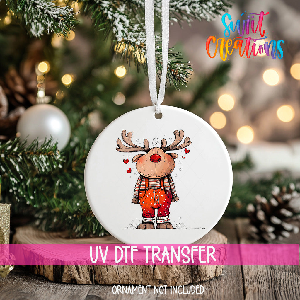 A white ornament with a cartoon reindeer design hangs on a Christmas tree, surrounded by pine branches and twinkling lights.