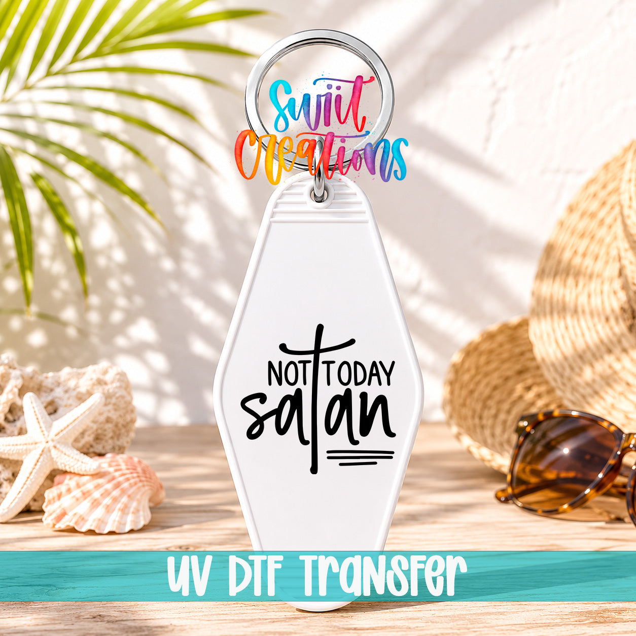 A keychain with a message that says "NOT TODAY SATAN" in a stylized font, placed on a wooden surface with sunglasses and a straw hat in the background.