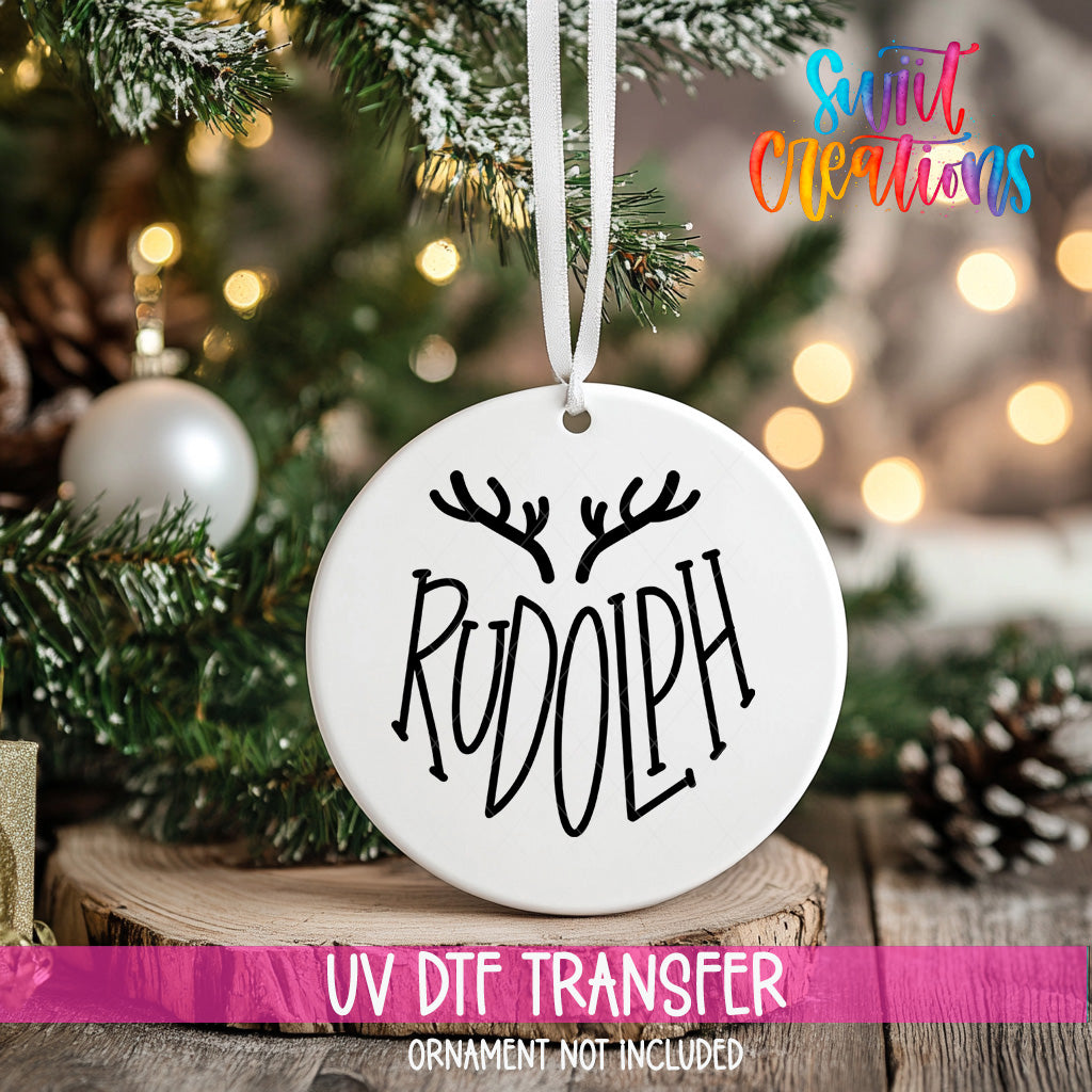 A white ornament with the word "RUDOLPH" printed on it, hanging from a Christmas tree.