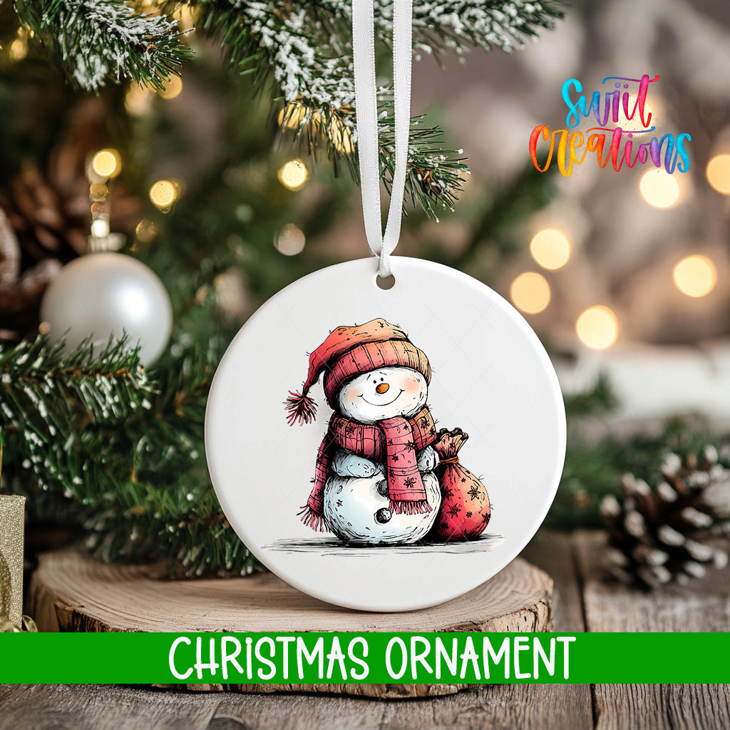 A white Christmas ornament featuring a snowman is hanging from a tree branch, with a blurred background of lights and greenery.