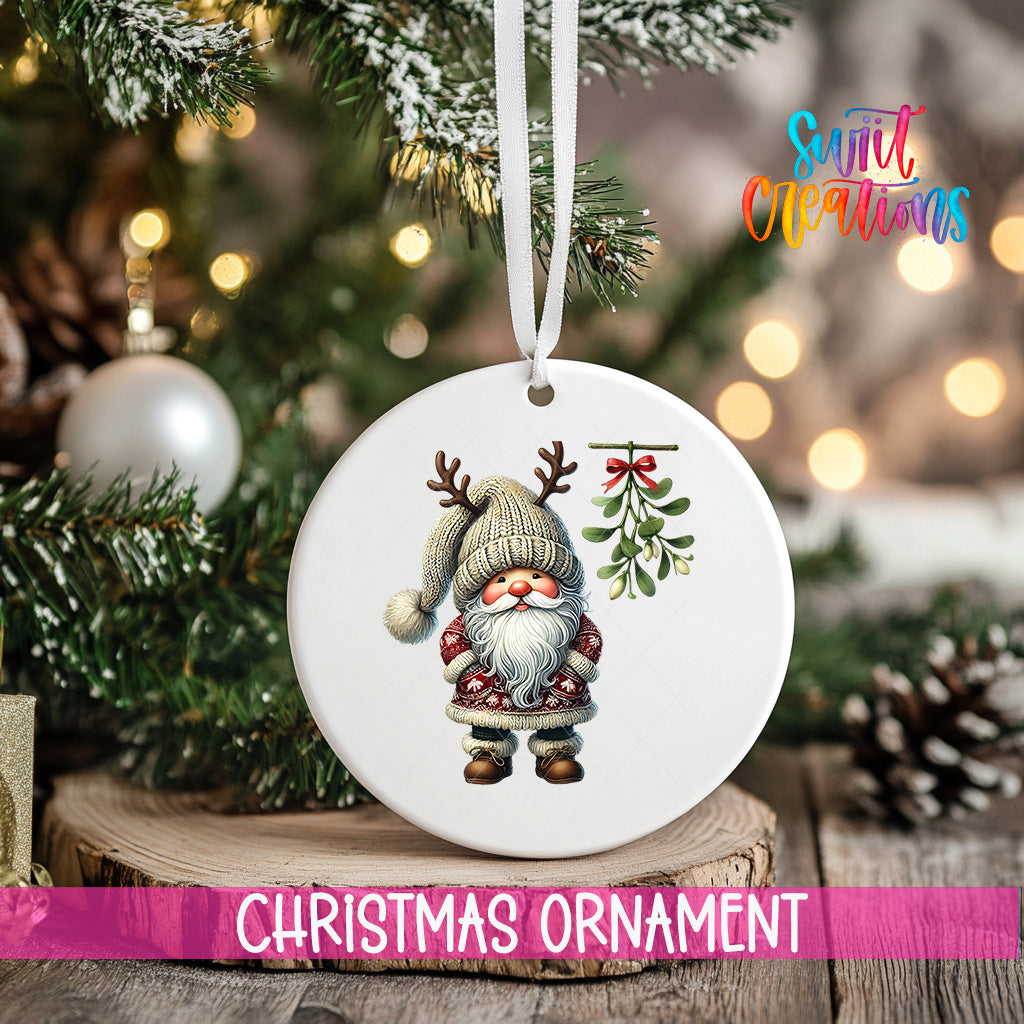 A white Christmas ornament featuring a gnome with antlers and a mistletoe is hanging from a tree branch.