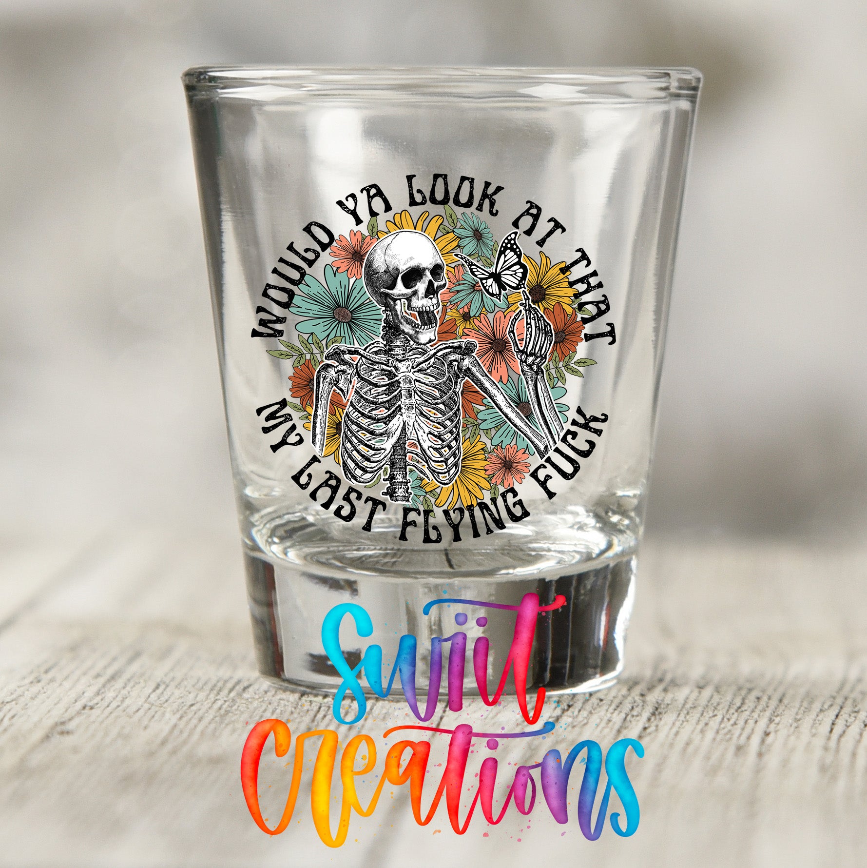 A shot glass with a skeleton and flowers design, along with the text "WOULD YA LOOK AT THAT MY LAST FLYING FUCK" printed on it.