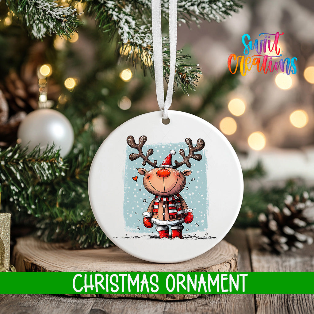 A white Christmas ornament featuring a cartoon reindeer with antlers and a scarf, hanging on a tree branch with a blurred background of lights and greenery.