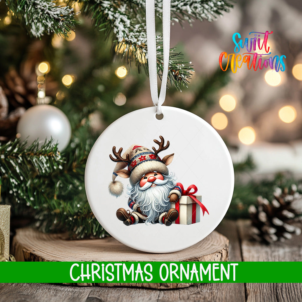 A white Christmas ornament featuring a smiling Santa Claus with antlers and a gift box, hanging on a tree branch with a blurred background of lights and greenery.