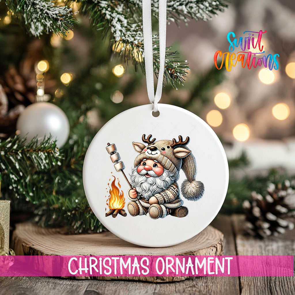 A white Christmas ornament featuring a cartoon Santa Claus holding a marshmallow over a campfire, with a pine tree and twinkling lights in the background.