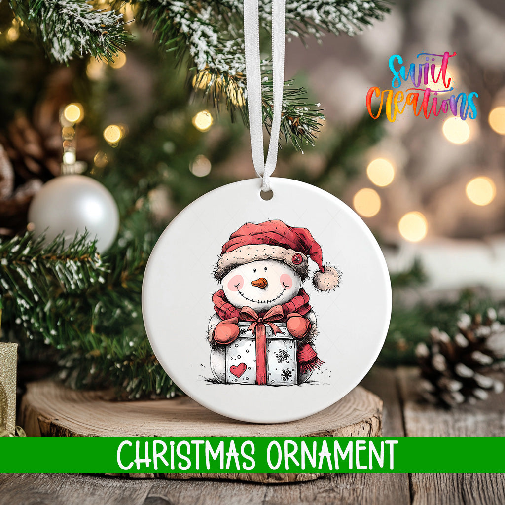 A white Christmas ornament featuring a smiling snowman holding a gift, hanging on a tree branch with a blurred background of lights and greenery.