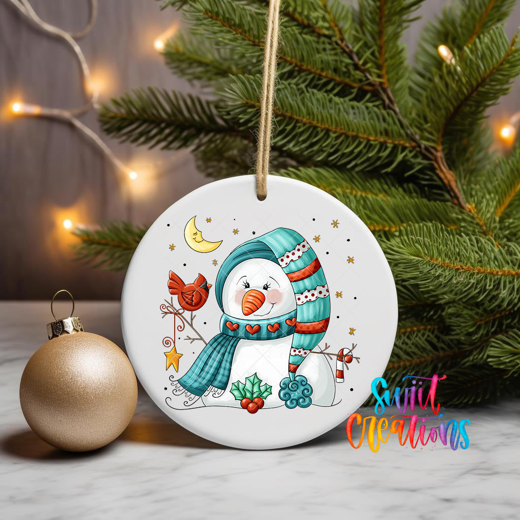 A white ceramic ornament featuring a snowman with a scarf and hat, surrounded by various holiday-themed elements such as a bird, a star, and a crescent moon. The ornament is hanging from a string, and there is a gold ornament next to it on a table. The background includes a Christmas tree and twinkling lights, creating a festive atmosphere.