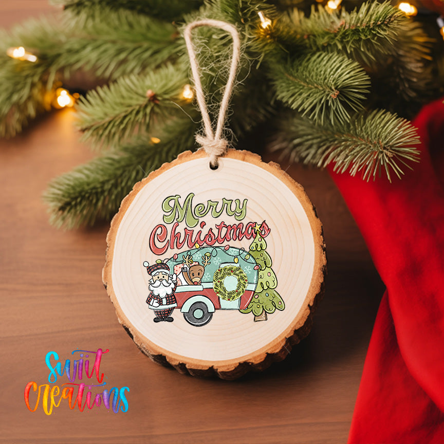 A wooden ornament with a Santa Claus and camper van design hangs on a Christmas tree, surrounded by greenery and twinkling lights.