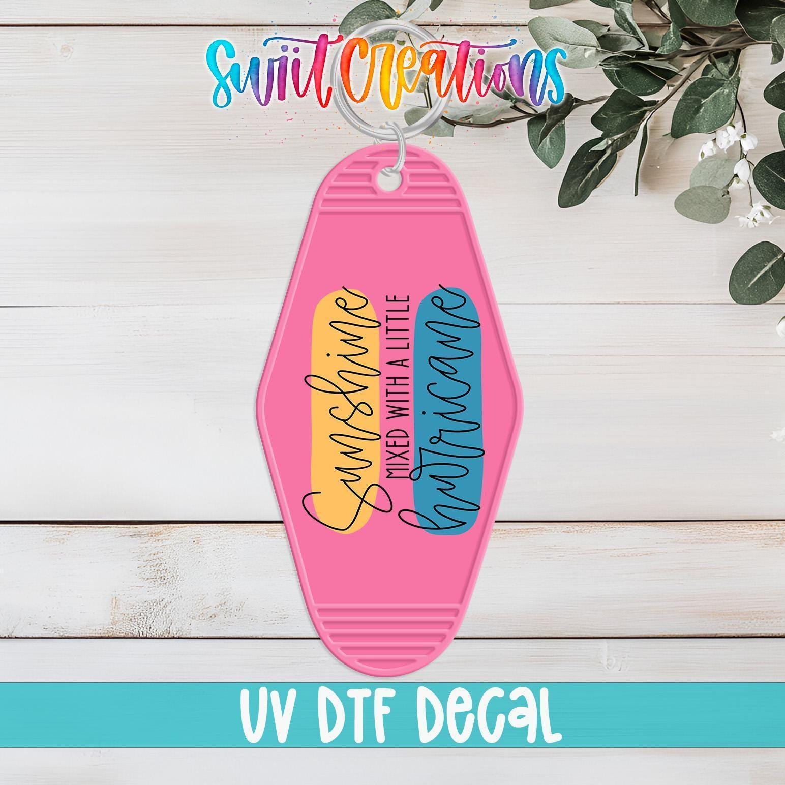 A pink keychain with a colorful design and text, placed on a wooden surface with greenery in the background.