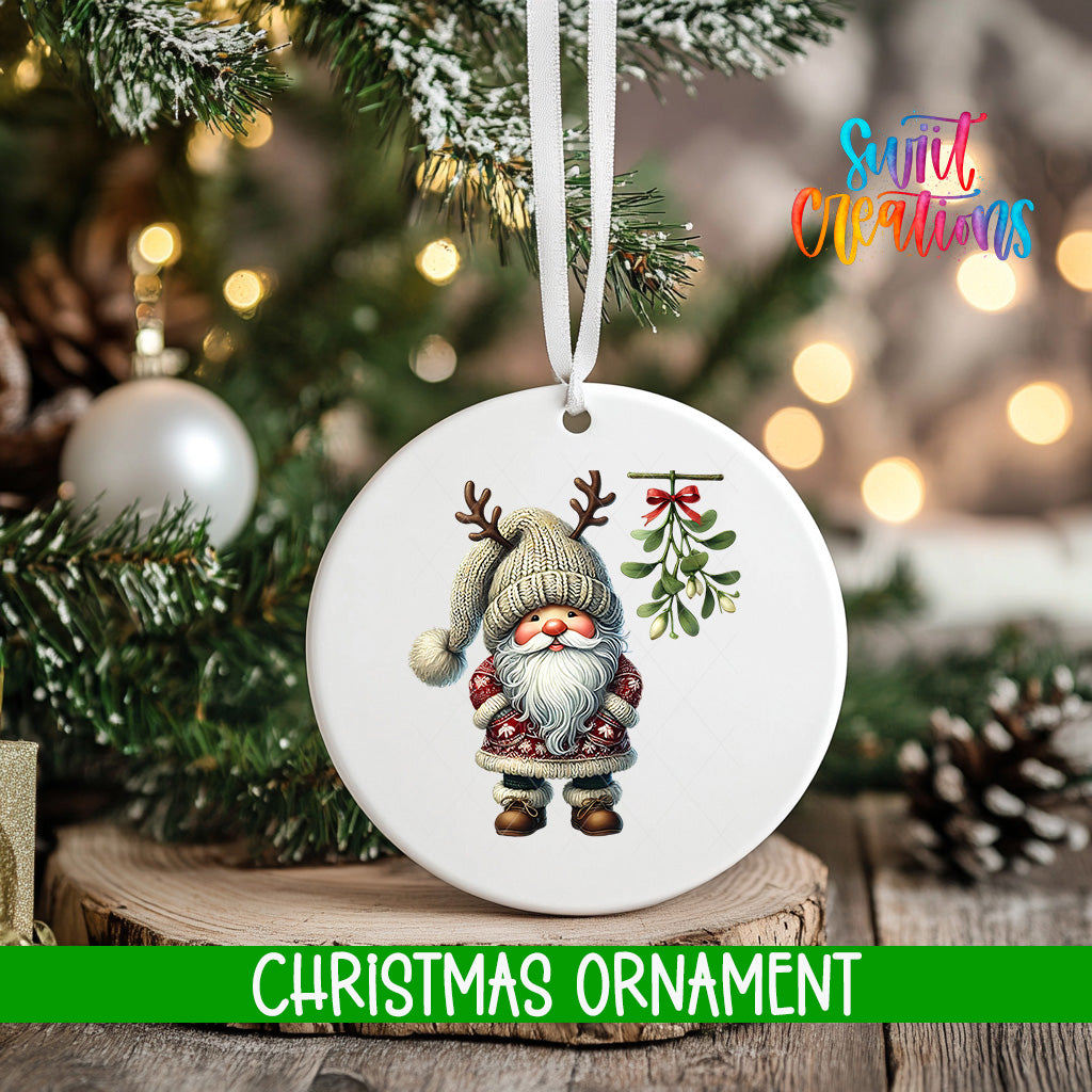 A white Christmas ornament featuring a gnome with antlers and a mistletoe is hanging on a tree branch.
