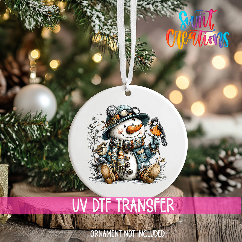 A white ornament with a snowman and a bird is hanging on a Christmas tree, with a pink ribbon around it.