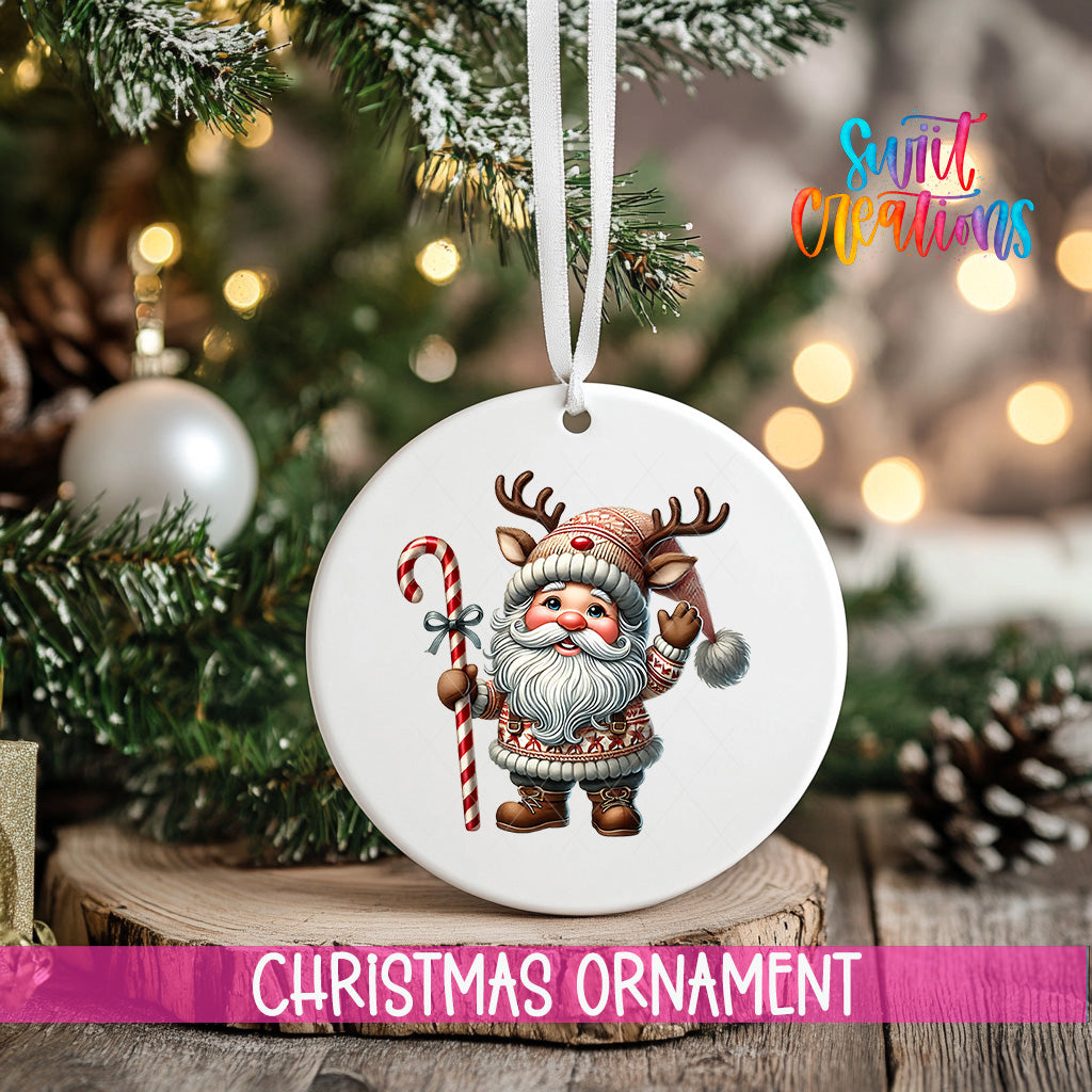 A white Christmas ornament featuring a cartoon Santa Claus holding a candy cane and reindeer antlers, hanging on a tree branch with a blurred background of lights and pine cones.