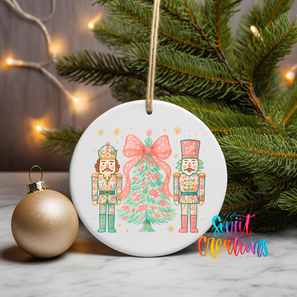 A white ceramic ornament featuring two nutcracker characters, one wearing a green outfit and the other in a pink outfit, standing in front of a Christmas tree. The ornament is hanging from a string, and there is a gold ornament and a Christmas tree visible in the background.