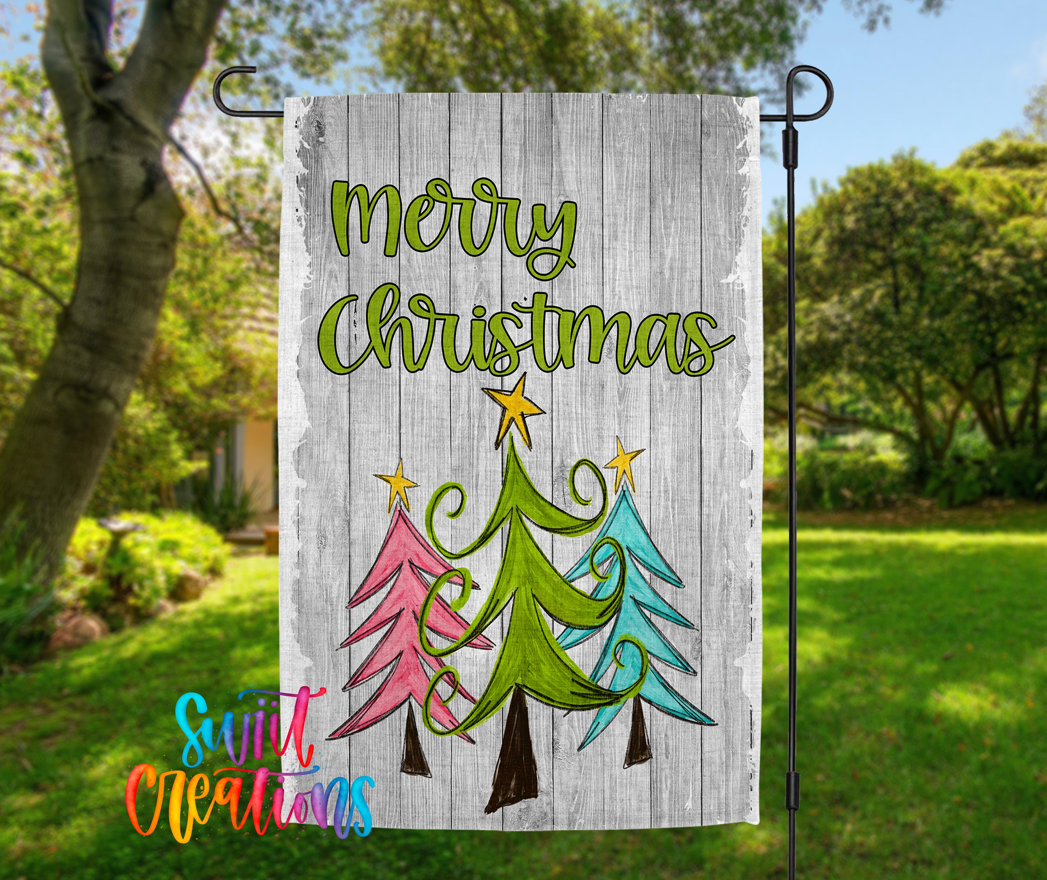 A colorful Christmas-themed flag is displayed in a garden setting, featuring a festive design of three stylized Christmas trees with a star at the top.